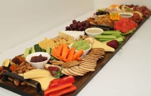A large grazing board with an assortment of cheeses, crackers, sliced vegetables, dips, fresh fruits, nuts, and cured meats, arranged neatly on a long wooden board. The display includes items like cucumber slices, carrot sticks, celery, watermelon, grapes, and a variety of spreads and dips, creating a colorful and appetizing spread.