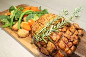 A roasted pork loin with crispy crackling, topped with a sprig of rosemary, served on a wooden board. The dish is accompanied by golden roast potatoes, steamed broccoli, green beans, and sliced carrots, creating a hearty and vibrant meal.