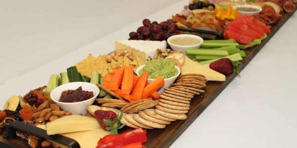 A large grazing board with an assortment of cheeses, crackers, sliced vegetables, dips, fresh fruits, nuts, and cured meats, arranged neatly on a long wooden board. The display includes items like cucumber slices, carrot sticks, celery, watermelon, grapes, and a variety of spreads and dips, creating a colorful and appetizing spread.