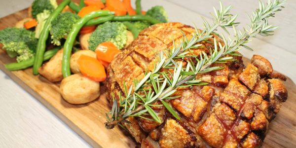 A roasted pork loin with crispy crackling, topped with a sprig of rosemary, served on a wooden board. The dish is accompanied by golden roast potatoes, steamed broccoli, green beans, and sliced carrots, creating a hearty and vibrant meal.